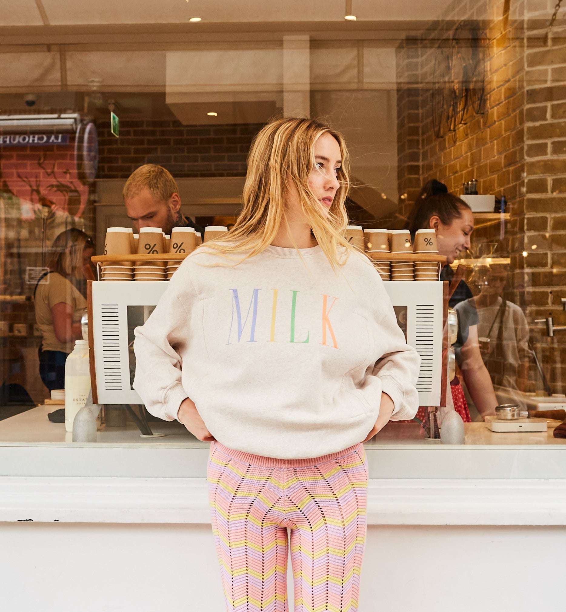 Blonde Frau trägt einen Tajinebanane Stillpullover, den man bei Mutterkleid kaufen kann. Auf dem Stillpullover ist MILK gestickt, alle vier Buchstaben sind in unterschiedlichen Farben (blau, gelb, grün und orange). Der Pullover ist beige und sie hat eine mehrfarbige Häkelhose kombiniert.