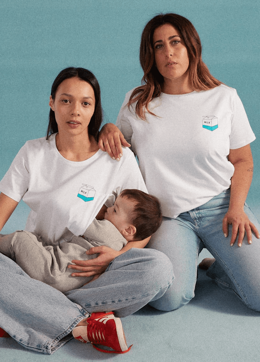 Zwei Frauen und ein Kind das gestillt wird. Beide Frauen tragen ein weißes Stillshirt mit kleinem Milchtüten Print auf der linken Brust. Dazu tragen die Frauen Blue Jeans und Sneaker