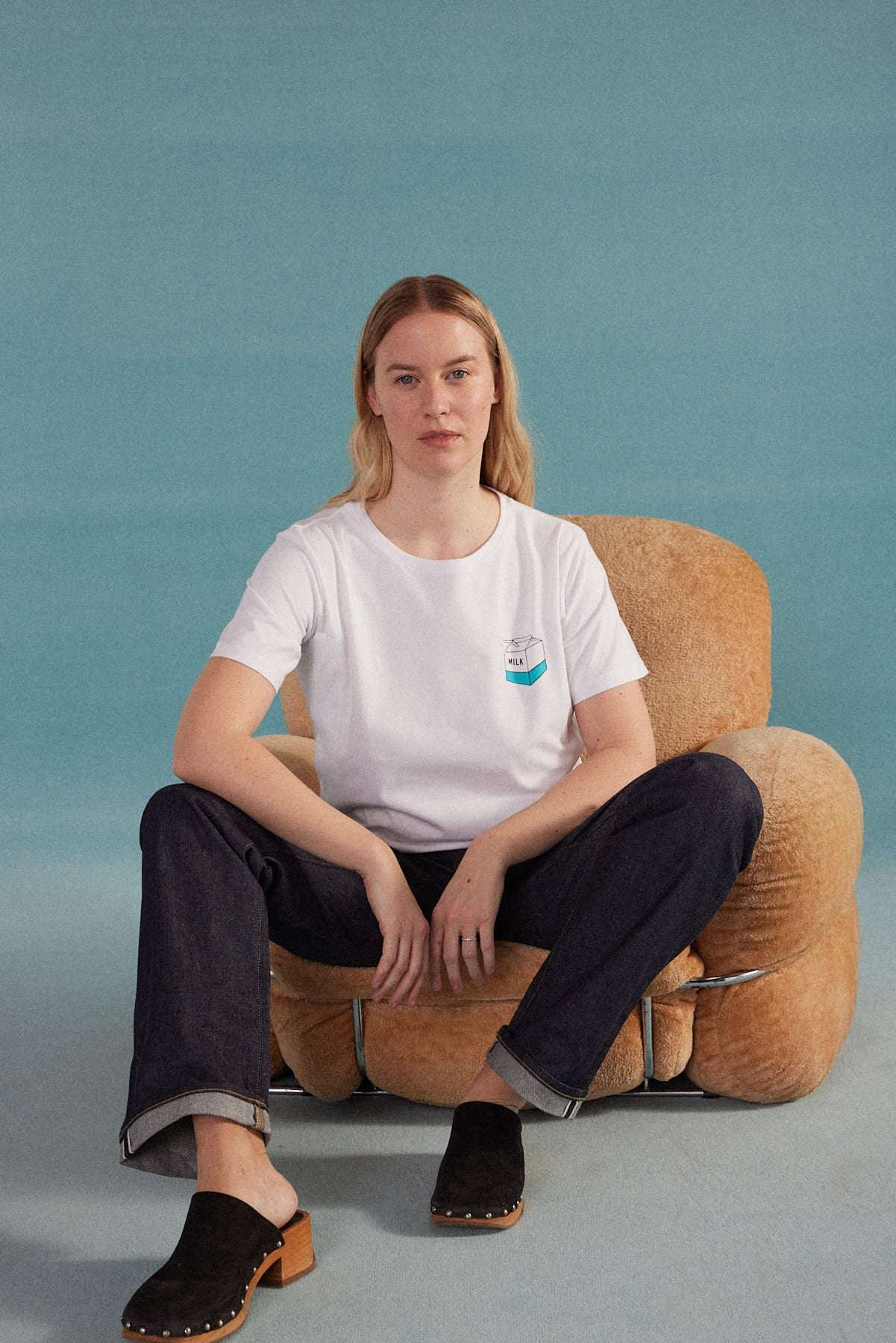 Frau sitzt auf einem Sessel und trägt ein weißes T-Shirt mit Stillzugang dazu schwarze Jeans und schwarze Clogs