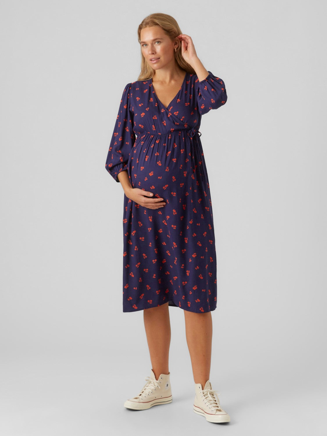 Blondes Model hat eine Hand an ihrem Ohr und zeigt frontal Mamalicious Umstandskleid mieten und online kaufen bei Mutterkleid Cherry Kirschen Print dunkelblau