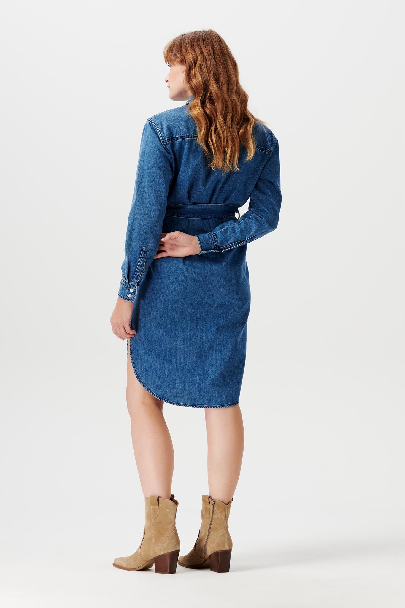 Im Mutterkleid Shop: Schwangeres Model zeigt lässiges Baumwoll Denim Umstandskleid von Noppies von hinten. Das Kleid hat lange Ärmel, die mit Knöpfen an den Handgelenken abschließen. Dazu stylt die bald Mama Cowboy Boots