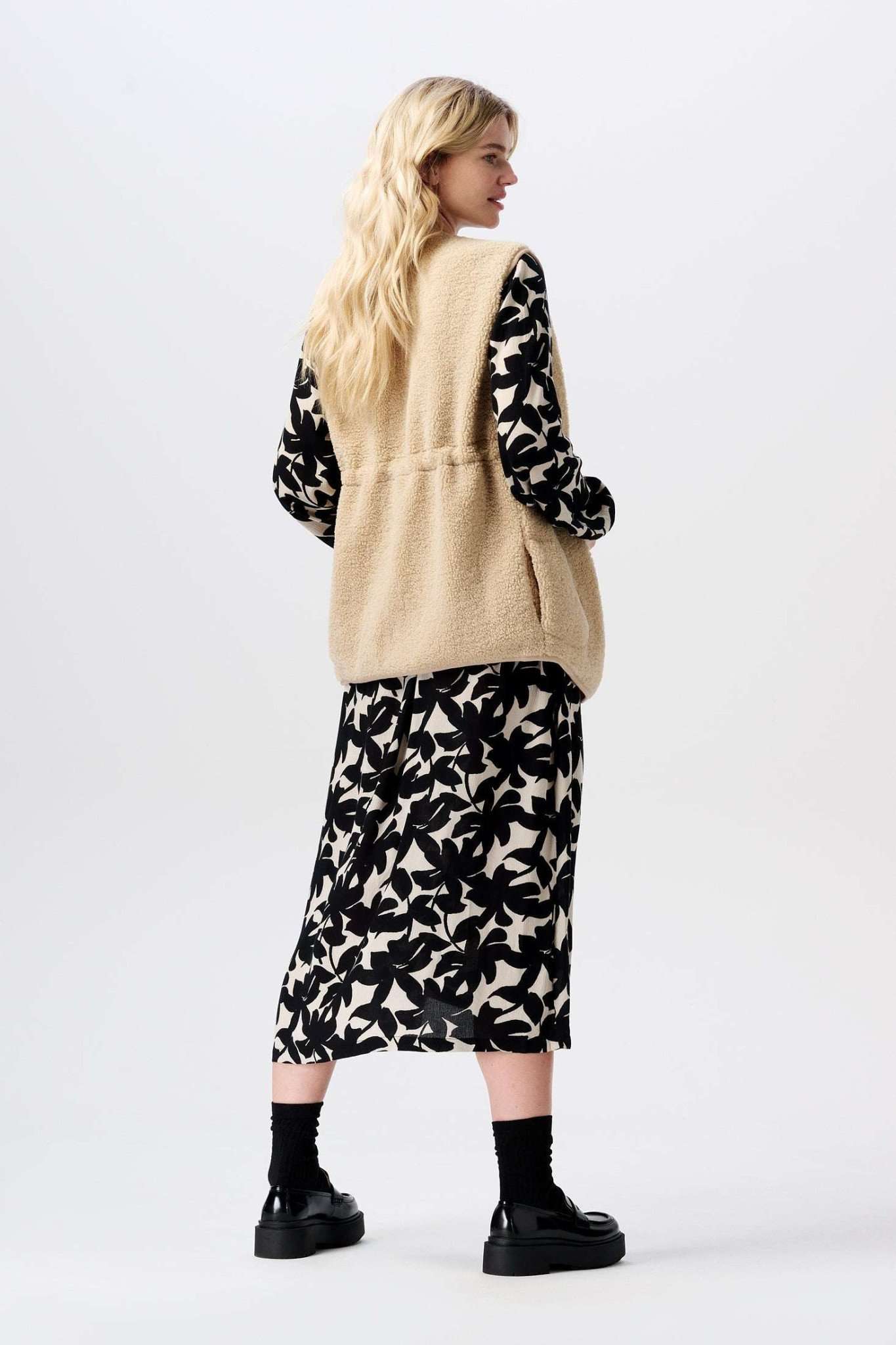 Schwangere Frau stylt die flauschige beige Teddy Umstandsweste von Noppies zu Midi Umstandskleid in schwarz und creme, erhältlich bei Mutterkleid.