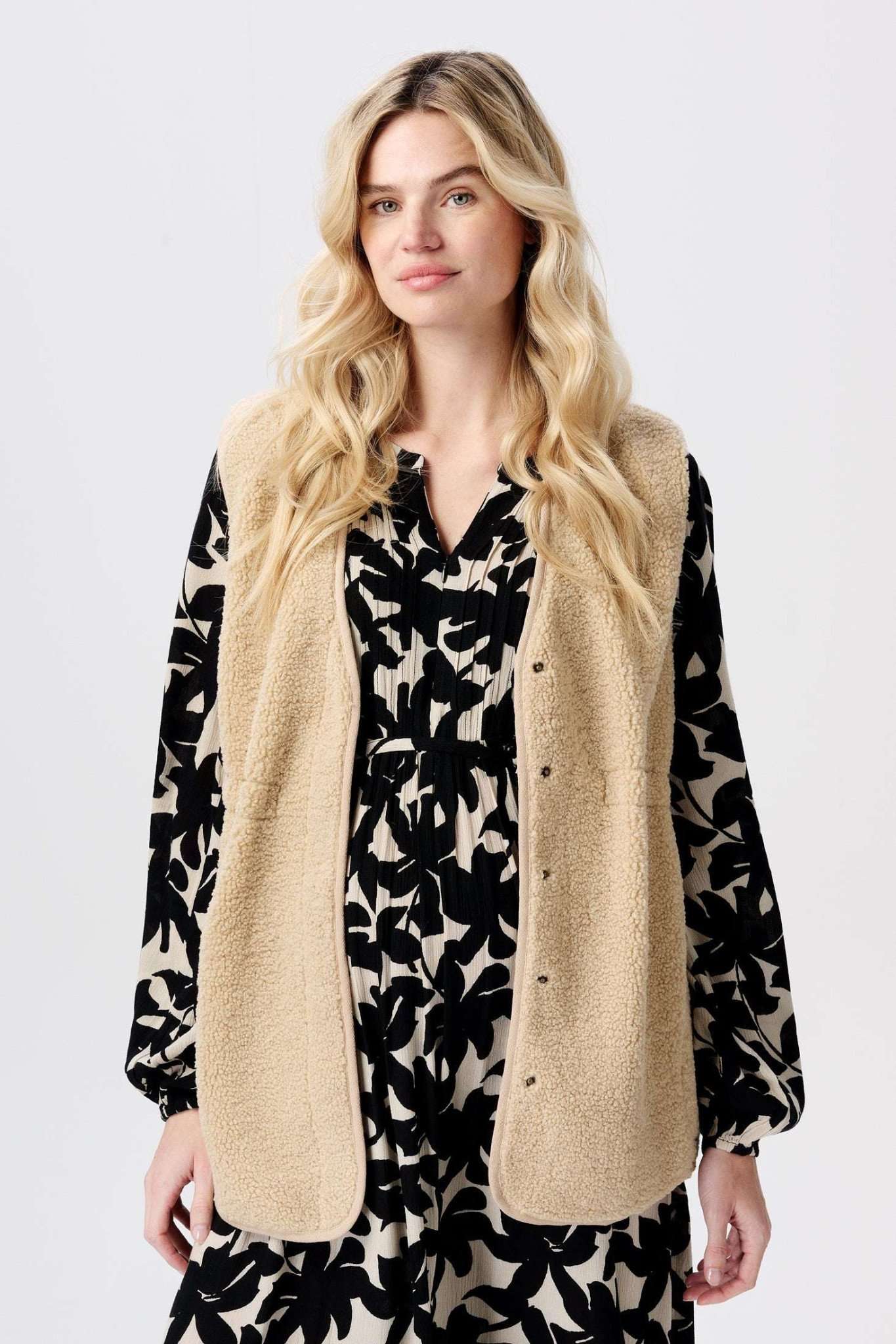 Das Model trägt ein florales Umstandskleid in schwarz creme und darüber eine beige Noppies Teddy Umstandsweste. Bei Mutterkleid kaufen oder mieten.