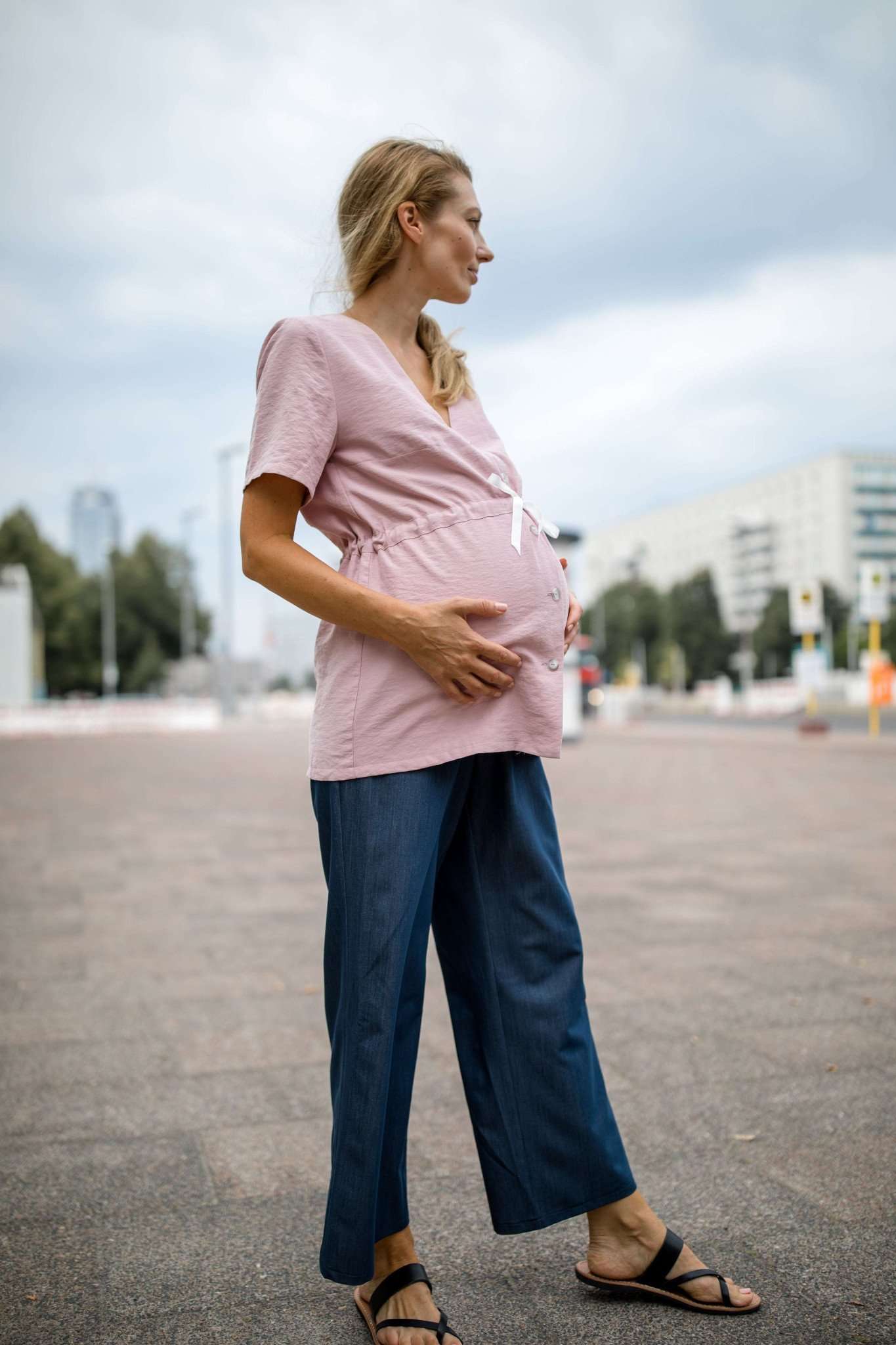 Bei Mutterkleid kaufen: Blondes Model steht auf der Straße und zeigt rosa Umstandsbluse im Profil.