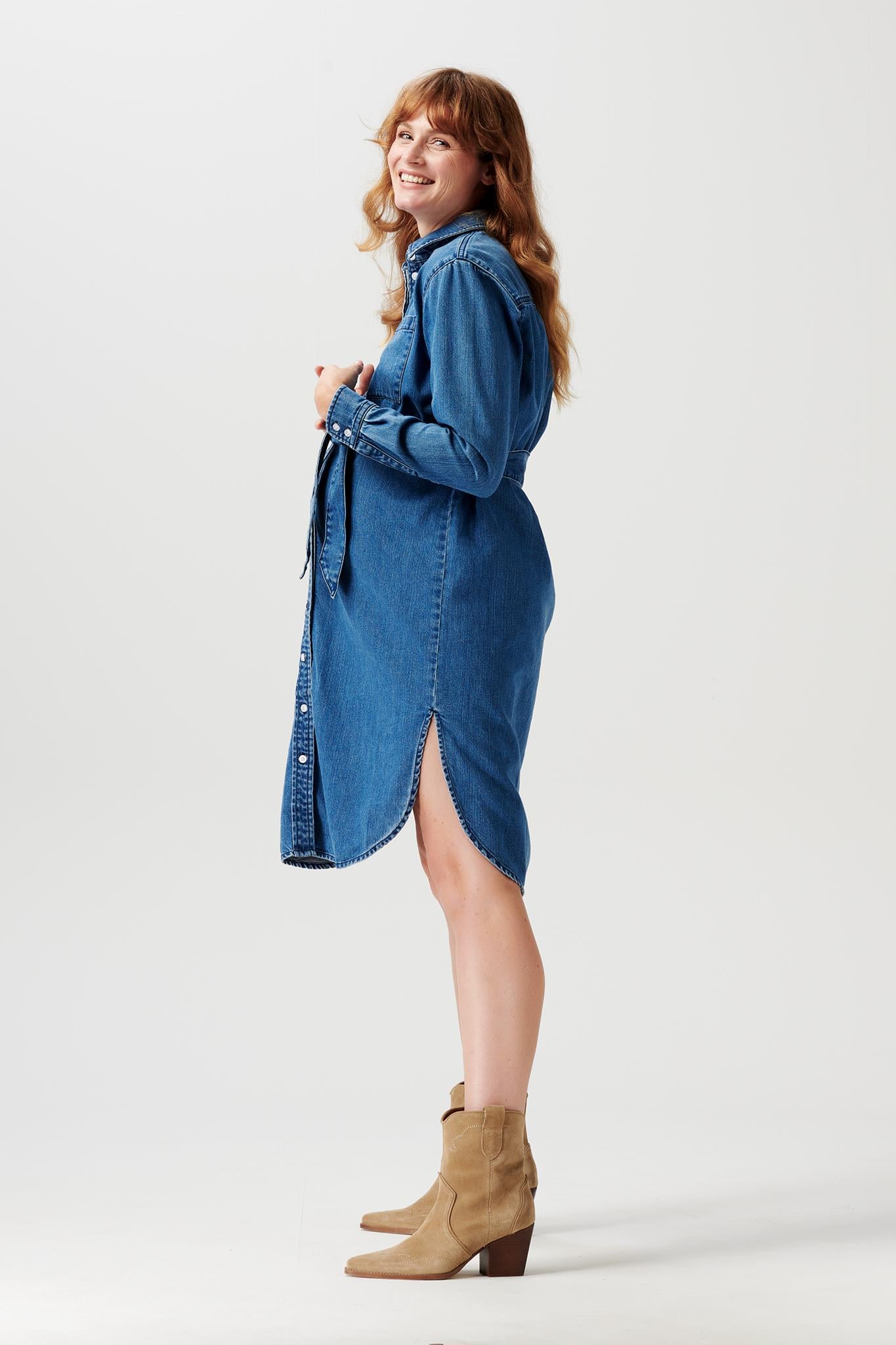 Lachende Schwangere im Profil fotografiert trägt Noppies Jeans Umstandskleid mit seitlichen Schlitzen und in Midi Länge, Hände liegen auf ihrem Babybauch. Im Mutterkleid Online Store kaufen oder mieten
