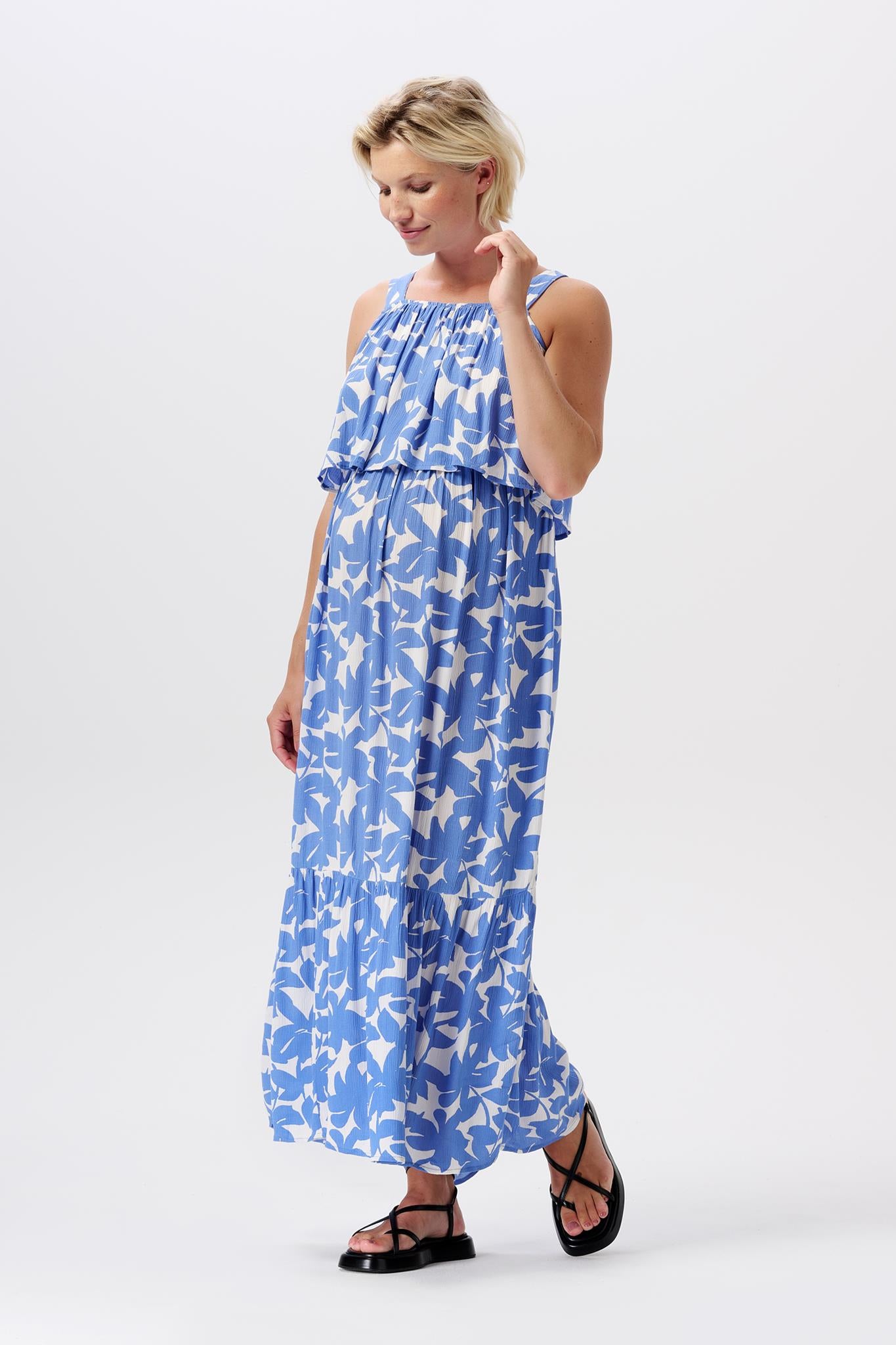 Modle mit kurzen blonden Haaren trägt Noppies Schwangerschaftskleid in Maxilänge mit blau weißen Floralprint aus leichter Viskose. Das Kleid umspielt die Knöchel. Volant über der Brust, unter dem sich der Stillzugang versteckt. Bei Mutterkleid.de einkaufen