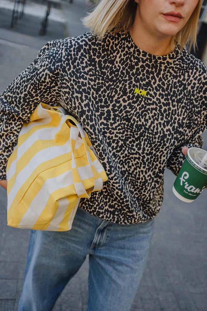 Frau spaziert mit Kaffee to Go auf der Straße. Sie trägt einen Baumwolle Pullover im Leo Print schwarz creme mit Neon MOM Print vorne. Dazu kombiniert eine blaue Jeans und eine gelb weiß gestreifte Shopping Bag