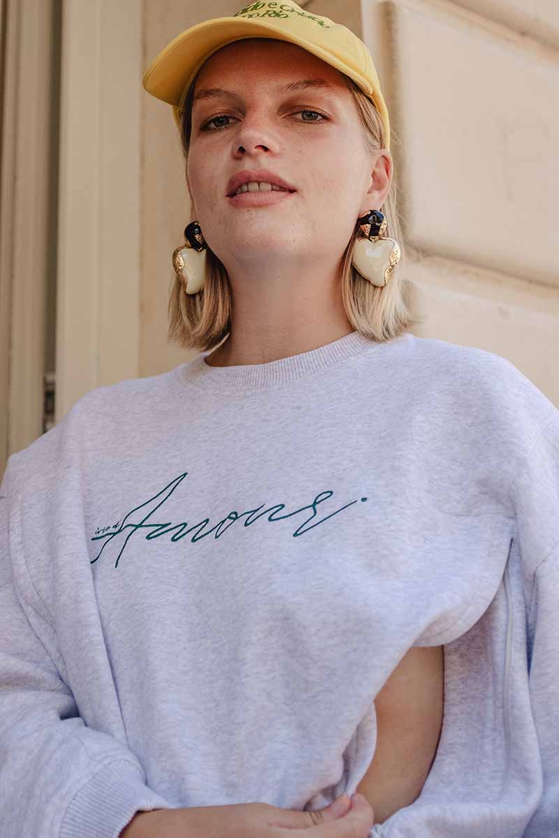 Stillende Frau trägt grauen Stillpullover mit seitlichem Reißverschluss für den Zugang zur Brust mit einer Amour Stickerei über der Brust. Mit gelber Cappie und großen Herzohrringen kombiniert