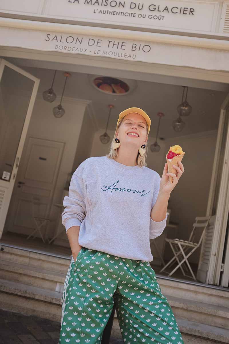 Lächelnde Frau mit einem Eis in der Hand. Sie trägt einen Baumwoll Pullover in grau mit grüner Amour Stickerei vorne. Dazu kombiniert eine gelbe Cappie, Herz Ohrringe und eine grüne adidas Satin Hose