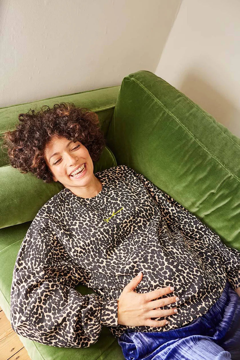 Model liegt lachend auf grünem Sofa  im Tajinebanane Umstandssweatshirt. Der Pullover ist im Leoprint und hat das Tajinebanane Logo in Neon vorne draufgestickt. Erhältlich bei Mutterkleid.de
