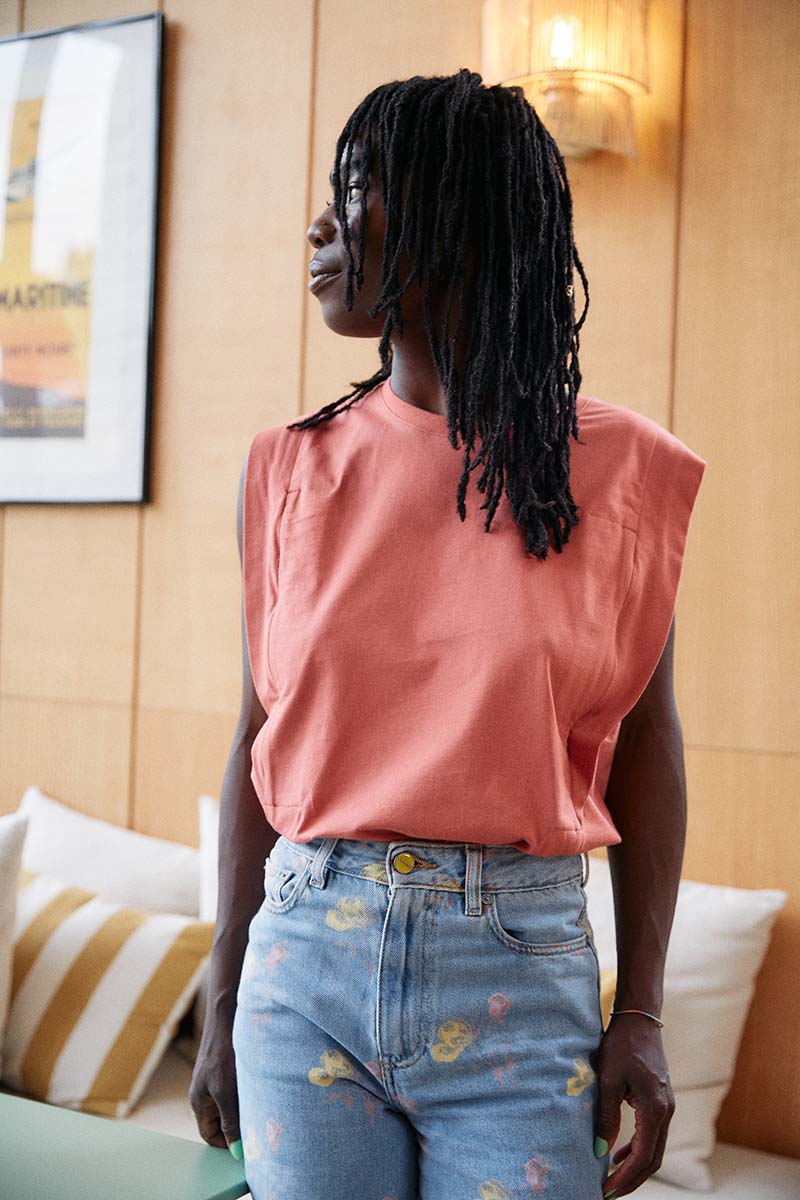 Frau steht im Wohnzimmer und trägt ein rotes T-Shirt mit überschnittenen Schultern das in einer blauen Jeans steckt