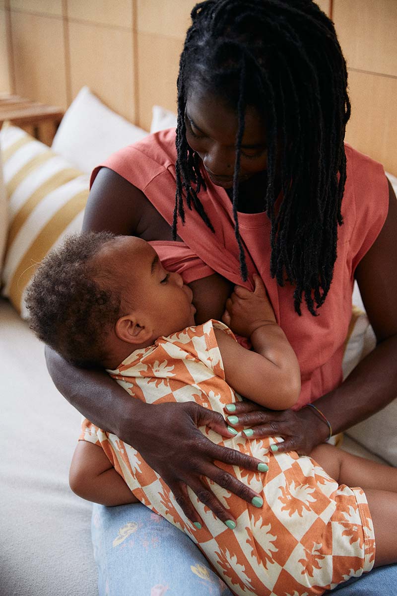 Stillende Mama mit Baby an der Brust. Das Stillshirt in rot hat seitliche Zugänge zur Brust