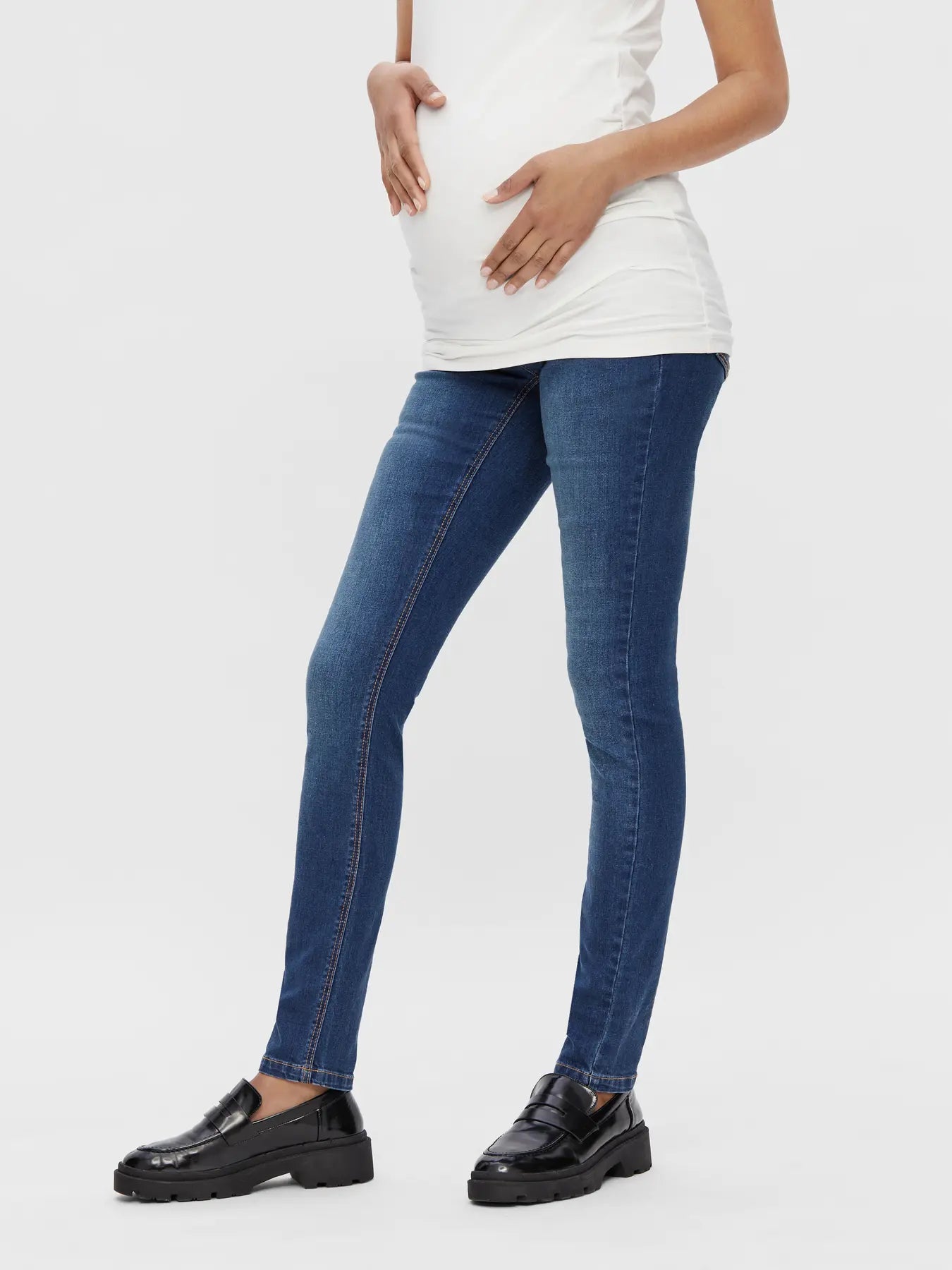 Seitliche Ansicht der blauen Mamalicious Umstandsjeans im Slim Fit - Model trägt weißes Umstands T-Shirt, Babybauch zeichnet sich ab