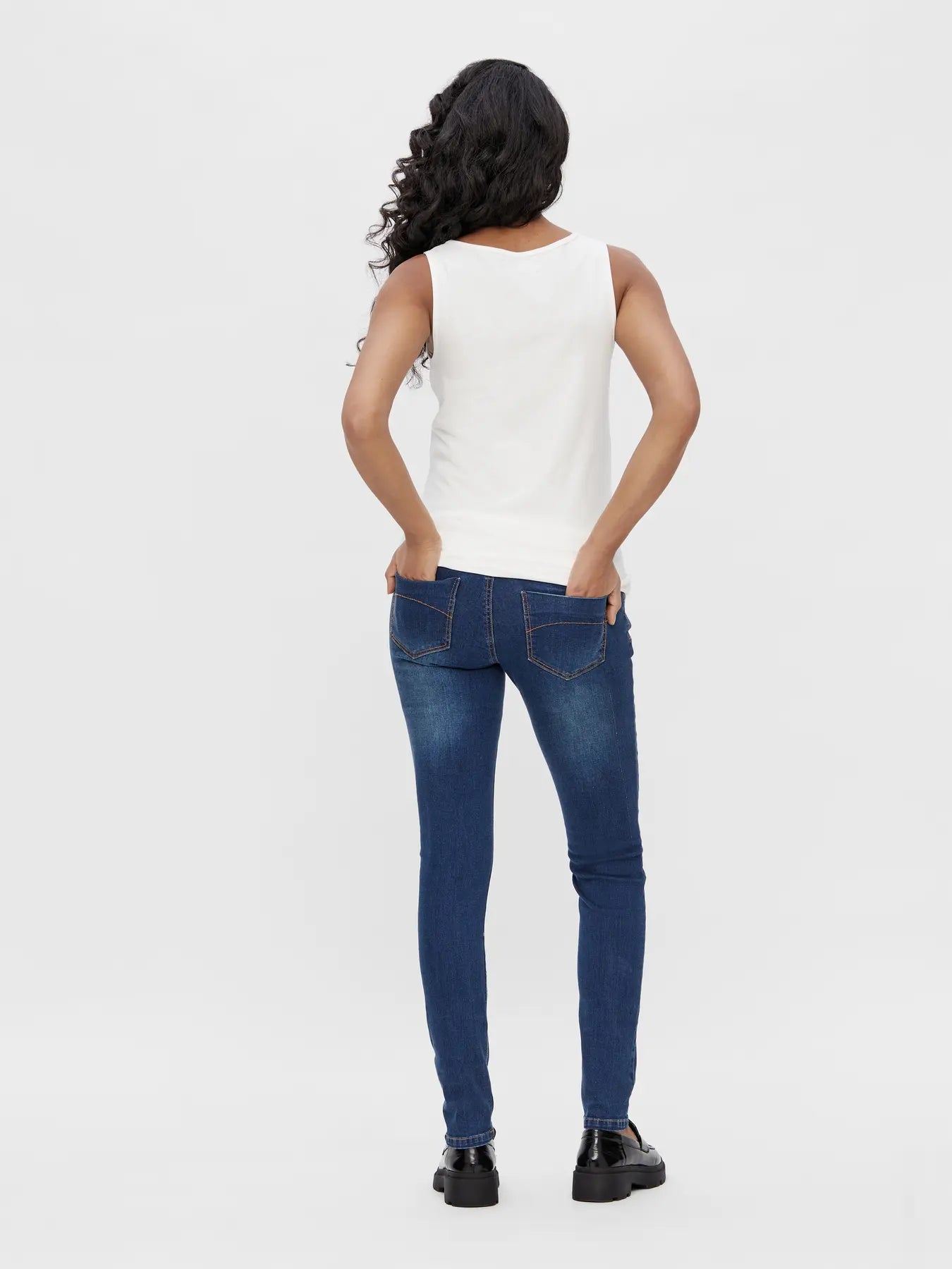 Rückansicht - Model trägt Slim Umstandsjeans aus blauem elastischem Denim - betonte Passform am Gesäß, beide Hände in den Gesäßtaschen