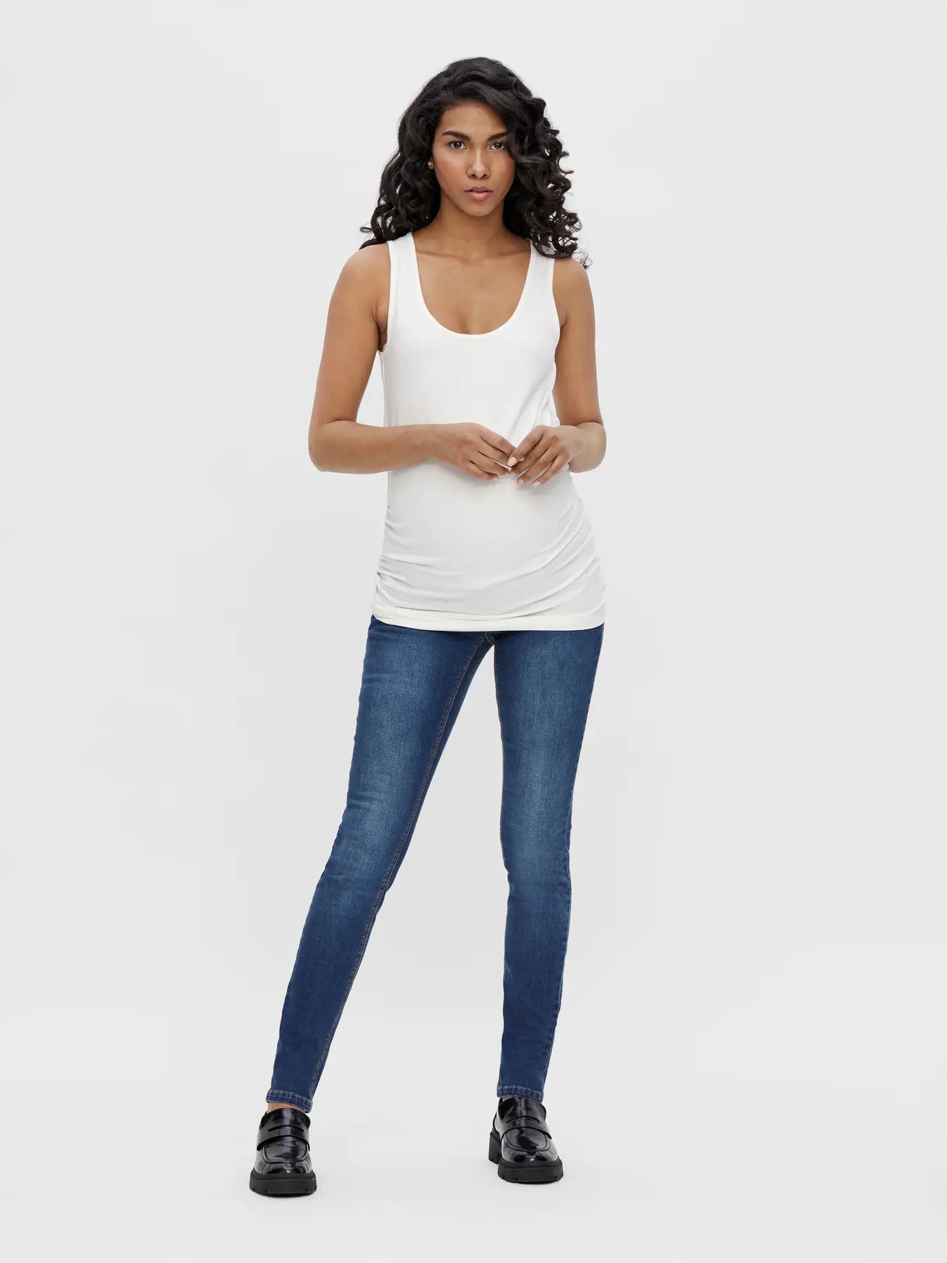 Ganzkörper Ansicht. Model schwanger trägt blaue Slim Umstandsjeans mit weißem Umstandsshirt, Babybauch sichtbar