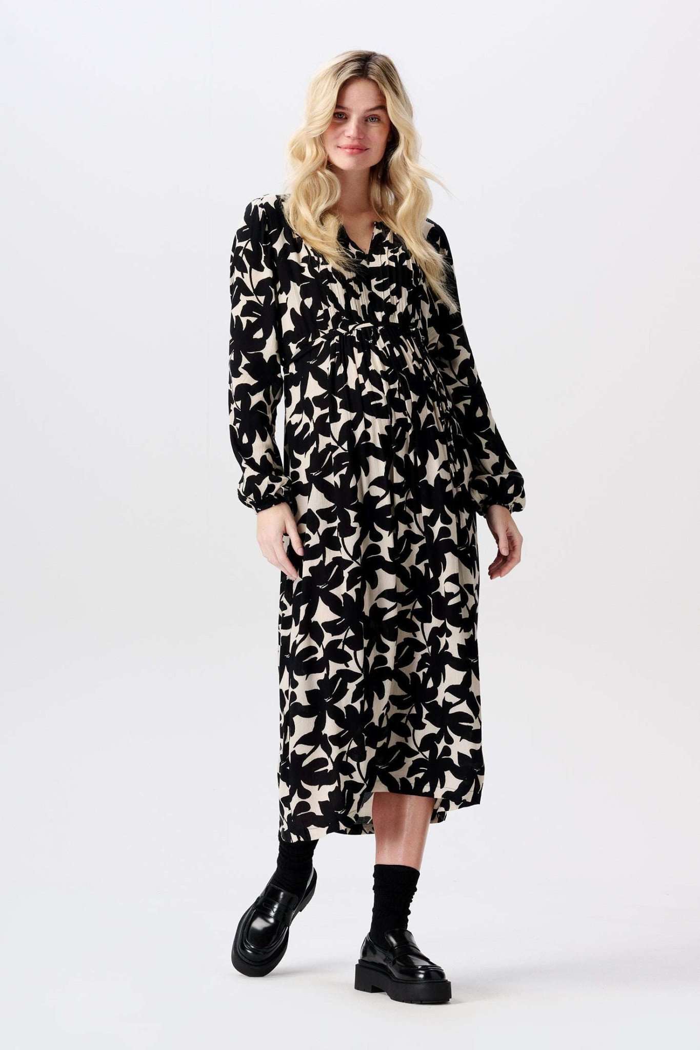 Schwangere blonde Frau trägt blumiges cremeweiß schwarzes Umstandskleid Stillkleid von Noppies in Midilänge mit langem Arm und dazu schwarze Loafer. Im Mutterkleid Shop online kaufen oder mieten.