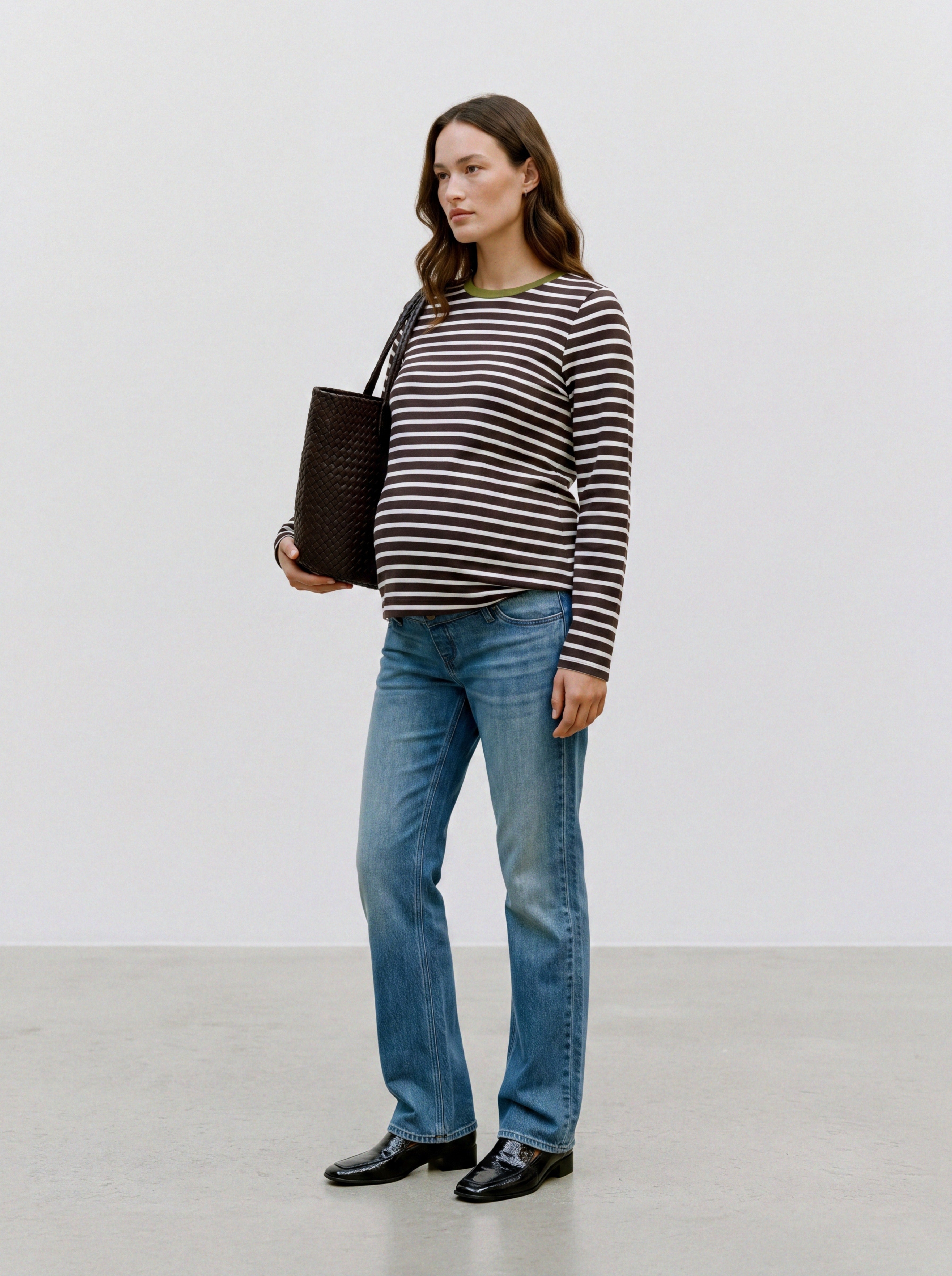 Schwangere Frau trägt gestreiftes Umstands-Longsleeve aus Baumwolle mit grünem Rundhalsausschnitt, Jeans und Tasche